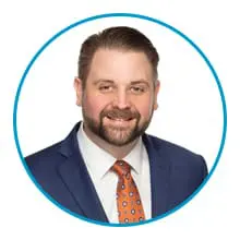 A man in a blue suit and an orange tie with a patterned design smiles against a white background within a blue circular border.