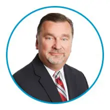 A man in a suit and tie is smiling inside a blue circular frame on a white background.