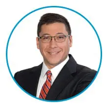 A man wearing glasses, a suit, and a red striped tie is smiling within a circular blue border.