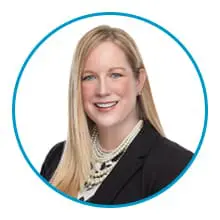 A person with long blonde hair is smiling, wearing a black blazer and a layered pearl necklace, set against a white background framed by a blue circle.