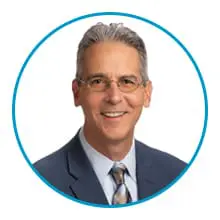 A smiling man with glasses is wearing a suit and tie, framed by a blue circle.