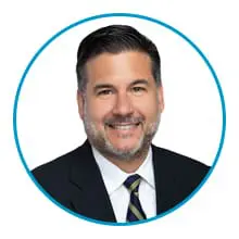 A smiling man with short dark hair and a beard is wearing a dark suit, white shirt, and striped tie inside a circular blue frame.