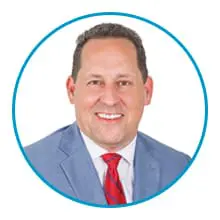 A person in a light blue suit, white shirt, and red-striped tie is smiling.