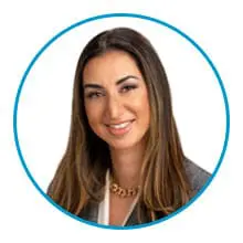 A woman with long brown hair is smiling, wearing a blazer and a gold necklace, framed in a blue circle.