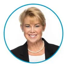 A professional-looking individual, wearing a suit jacket, pearl necklace, and smiling within a blue circular frame.