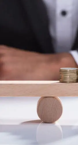 Close-up Of A Businessperson's Hand Balancing Stacked Coins On Wooden Seesaw With Finger Over Desk