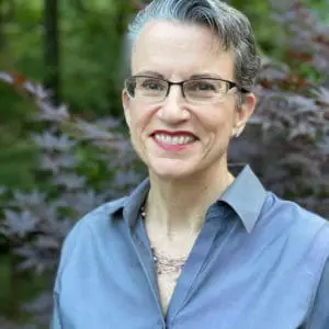 A person with short gray hair and glasses is smiling in an outdoor setting with trees in the background.