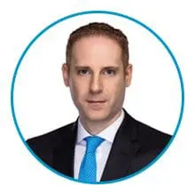 A man with short, reddish-brown hair is wearing a black suit, white shirt, and blue tie, framed within a blue circle against a white background.