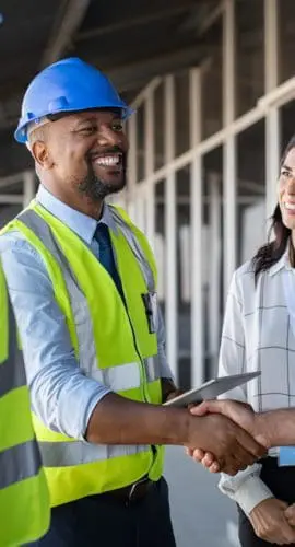 Smiling engineer shaking hands at construction site with happy architect. Handshake between cheerful african construction manager with businessman at bulding site. Team of workers with architects and contractor conclude an agreement with safety uniform.