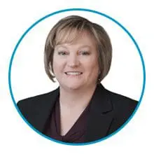 A person with short, light brown hair is smiling while wearing a dark blazer.