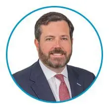 A man with a beard, wearing a suit and a red tie, is inside a blue circular frame on a white background.