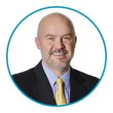 A smiling bald man wearing a suit with a yellow tie inside a blue circular frame.