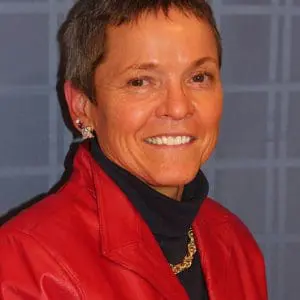 A person with short hair, wearing a red jacket and a black turtleneck, smiles in front of a blue background.