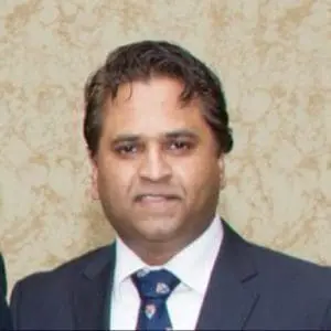 A man in a suit and tie stands in front of a patterned beige wall.