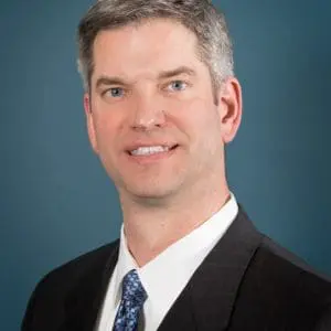 A man wearing a dark suit, white shirt, and blue patterned tie is posing against a blue background.