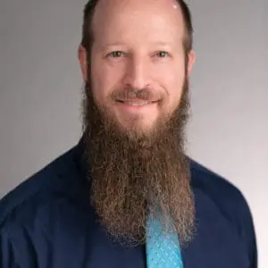 A man with a long beard is smiling and wearing a dark shirt with a light blue tie.