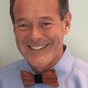A smiling man is wearing a light purple shirt and a wooden bowtie.
