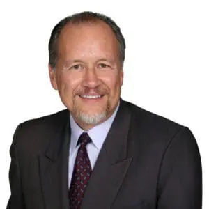 A man with short hair and a beard is smiling while wearing a dark suit, white shirt, and patterned tie against a white background.