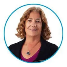 A person with shoulder-length curly hair wearing a black jacket and a necklace smiles against a white background enclosed in a blue circle.