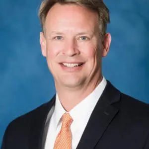 A smiling person in a suit with a white shirt and an orange tie is set against a blue background.