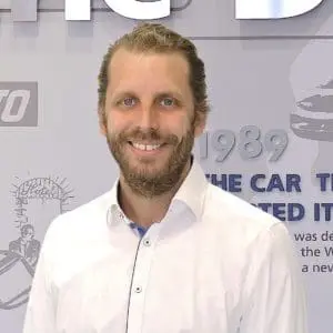 A man with a beard and tied-back hair is smiling and wearing a white dress shirt in front of a backdrop with text and logos.