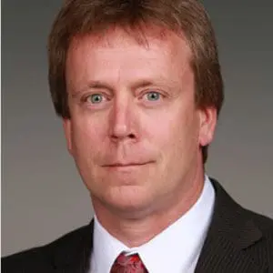A person with short, light brown hair wearing a suit and a red tie against a plain background.
