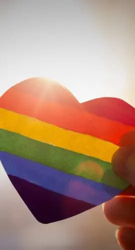 hand holds a heart painted like a LGBT flag, silhouetted against sun