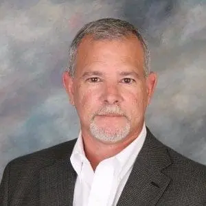 A man with gray hair and a neatly trimmed beard is wearing a dark suit jacket and white shirt against a cloudy backdrop.