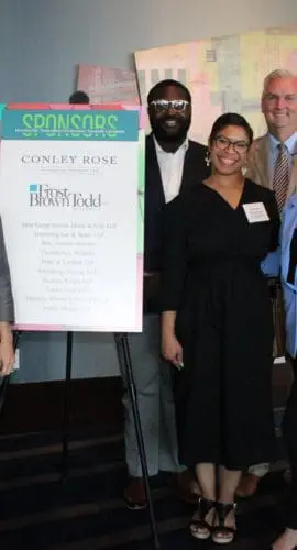 A group of people is standing and smiling around an easel displaying an event sign with sponsorship details.