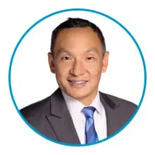 A person in formal attire, wearing a gray suit, white shirt, and blue striped tie, is facing forward and smiling.