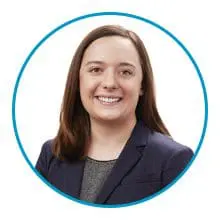 A person with shoulder-length brown hair is smiling and wearing a dark blazer over a gray top, encircled by a blue line.