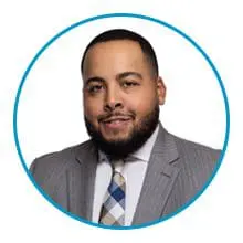 A person with a beard and short hair is wearing a gray suit with a patterned tie, framed within a blue circle.