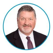 A person with gray hair and a beard is wearing a suit and tie, framed by a blue circle.