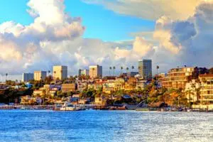 A coastal cityscape features modern buildings against a backdrop of a cloudy sky and a vibrant blue ocean in the foreground.