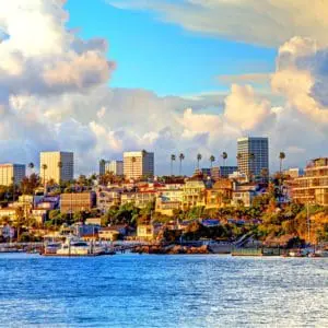 A coastal cityscape features modern buildings against a backdrop of a cloudy sky and a vibrant blue ocean in the foreground.