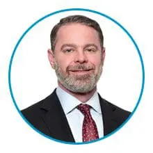 A man with a beard and short hair, wearing a suit and a red tie, is shown in a circular frame.