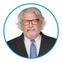 An older man with gray hair and glasses is dressed in a suit and tie, looking directly at the camera within a circular frame.