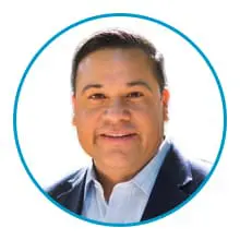 A smiling person wearing a light-colored shirt and a dark blazer within a circular blue frame.