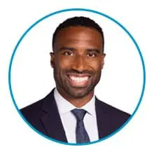 A man wearing a dark suit, white shirt, and dotted tie is smiling in a headshot with a blue circular border.