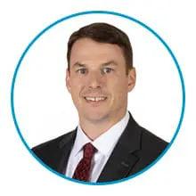 A man with short brown hair is wearing a dark suit, white shirt, and red tie, and is encircled by a blue border.