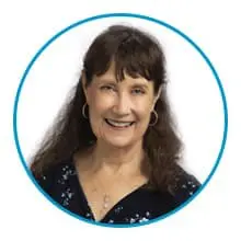 A woman with long brown hair and hoop earrings is smiling and wearing a dark blue floral top and a necklace.