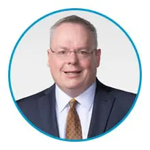 A man wearing glasses, a suit, and an orange tie stands against a light background within a blue circular border.