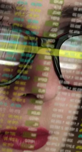 Stock photo of a beautiful young woman’s face very close up. She’s looking at a complicated set of holographic, see-thru numbers. The numbers are also reflected in her spectacles.