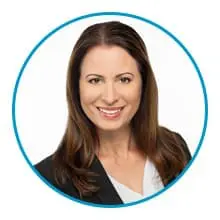 A woman with long brown hair wearing a black blazer and white top is smiling at the camera within a blue circular border.