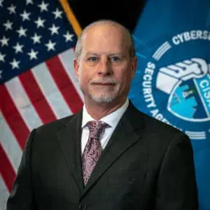 A man in a suit stands in front of an American flag and a cybersecurity agency's flag.