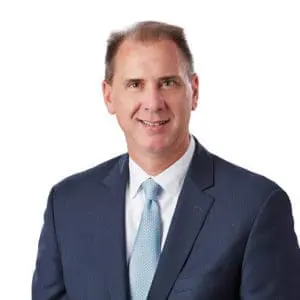 A man wearing a blue suit and light blue tie smiles against a plain white background.