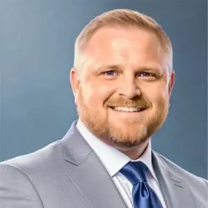 A smiling man with a beard is wearing a gray suit and a blue tie.