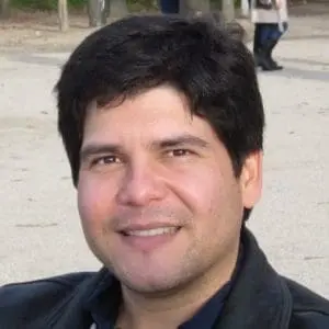 A person with short, dark hair is smiling outdoors, wearing a dark jacket.