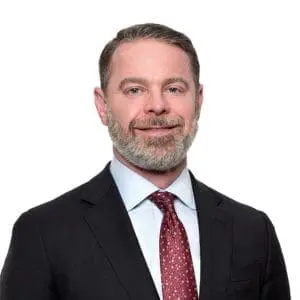 A man with a beard is wearing a black suit, light blue shirt, and red patterned tie, looking at the camera with a neutral expression.