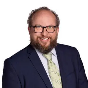 A person with glasses, a beard, and curly hair is wearing a dark suit jacket, light blue dress shirt, and a light green tie with circular patterns.